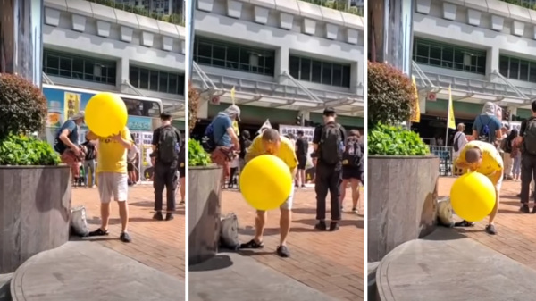 阿雄站在地上拿起一个偌大的黄气球，用力使劲地吹，每吹一次，身体也随之向前低俯。（图片来源：看中国视频截图）