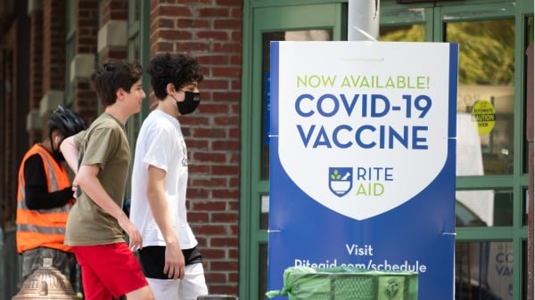 美國紐約市街頭走過COVID-19疫苗接種指示牌的兩位少年，一人帶口罩，一人不帶，前者情緒似乎不高，而後者在微笑。